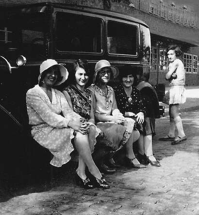 Four ladies sitting on the footboard of a car.